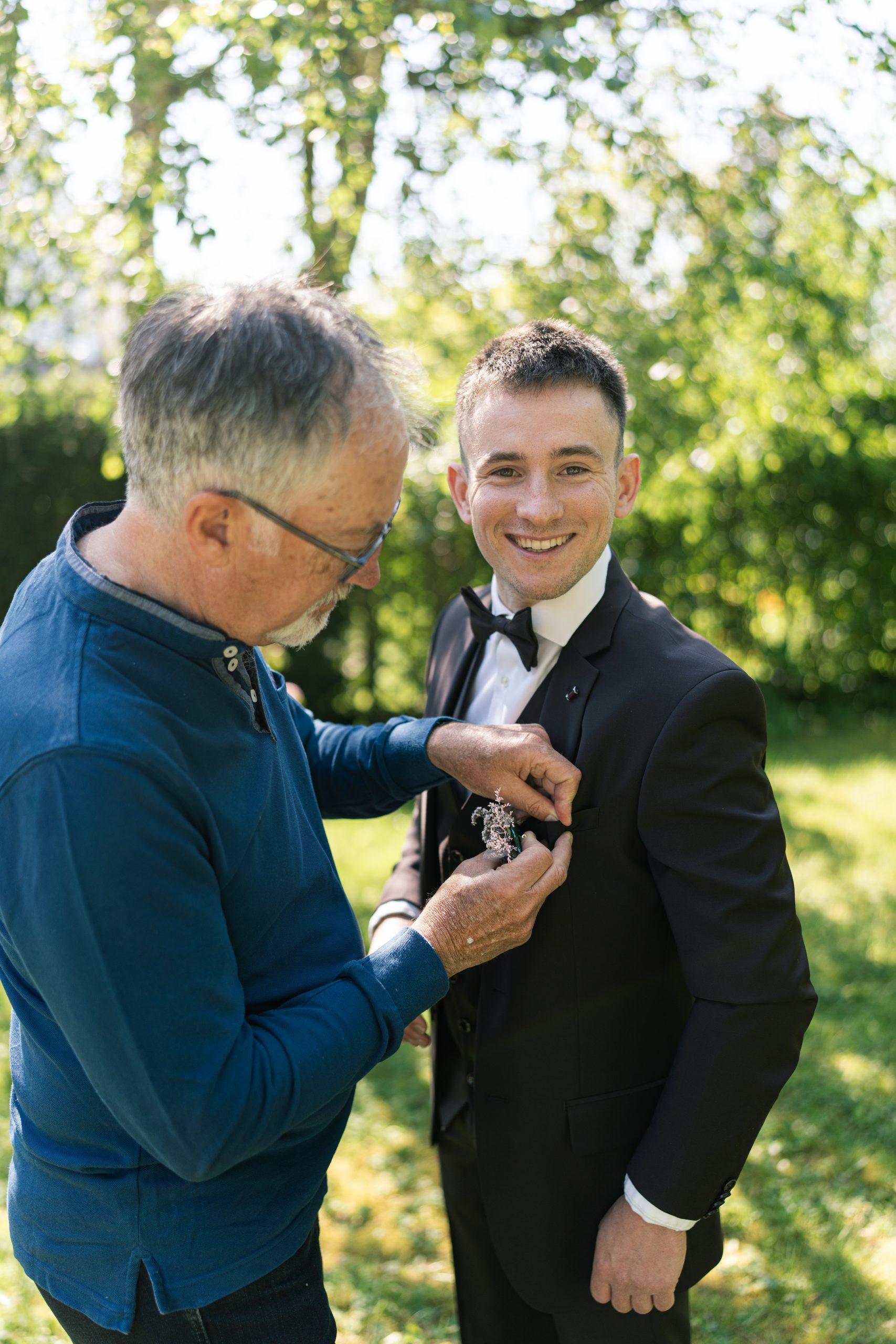 Un proche fixe la boutonnière fleurie de Théo sur son costume de mariage