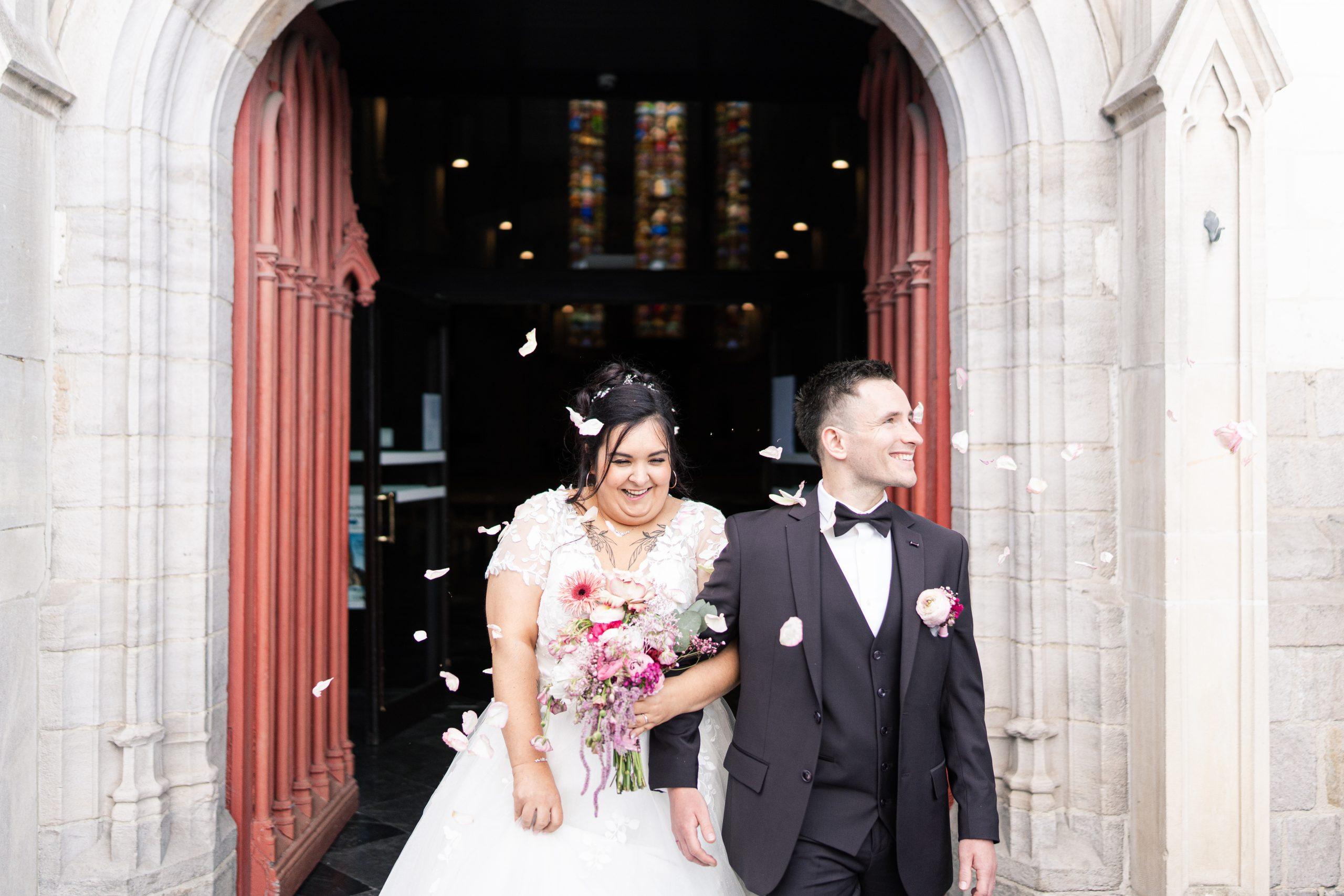 Émilie et Théo sourient sous les pétales de fleurs à la sortie de l’église
